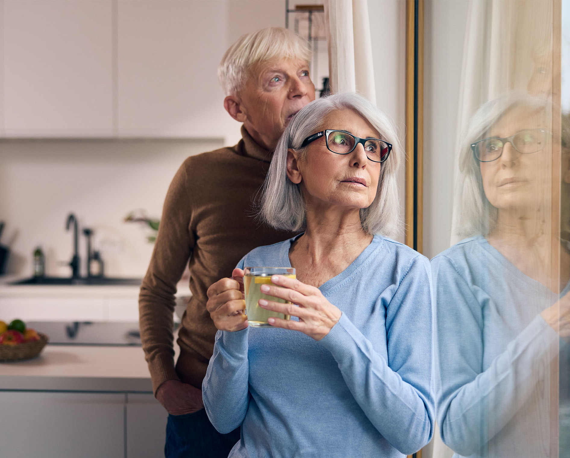 Älteres Paar blickt nachdenklich an einem Fenster in die Ferne. Die Frau hält eine Tasse Tee.